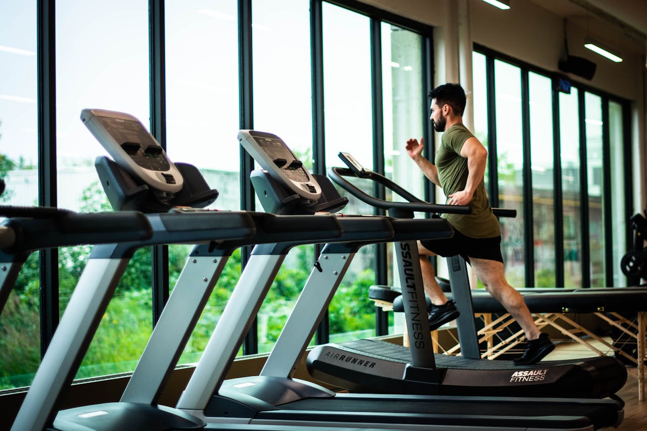 About Young man workouts on treadmill in modern gym with large windows and natural light.